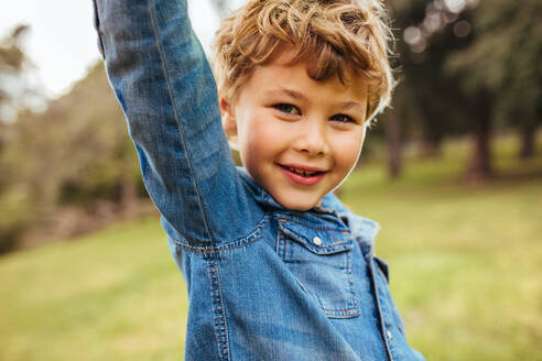 Nahaufnahme eines niedlichen kleinen Jungen, der mit erhobener Hand im Freien steht und ein Jeanshemd trägt und in die Kamera schaut. - JLPSF15841