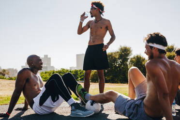 Fußballspieler, die während einer Pause auf dem Boden sitzen und sich unterhalten. Müder Sportler, der nach einem Fußballspiel an einem sonnigen Tag Wasser trinkt. - JLPSF15847