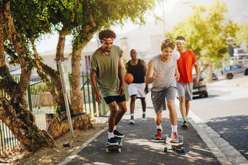 Zwei Männer skaten auf einem Skateboard auf einem Bürgersteig, während ihre Freunde sie anfeuern. Basketballer, die auf einem Bürgersteig mit Skateboards laufen. - JLPSF15851