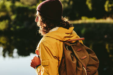 Nahaufnahme eines Mannes in Jacke, der im Freien spazieren geht und einen Rucksack trägt. Mann, der mit Wintermütze und Jacke an einem See spazieren geht. - JLPSF15860