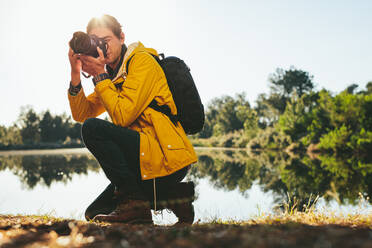 Fotograf, der an einem sonnigen Tag an einem See sitzend die Natur fotografiert. Ein Reisender, der die Schönheit der Natur in einer ländlichen Umgebung mit der Kamera festhält. - JLPSF15865