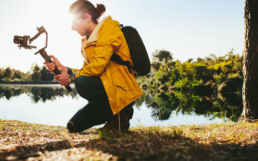 Ein Mann, der mit modernsten technischen Hilfsmitteln Fotos von der Natur schießt. Ein Reisender sitzt an einem See und macht ein Foto mit einer DSLR-Kamera, die auf einem handgehaltenen Gimbal montiert ist. - JLPSF15866