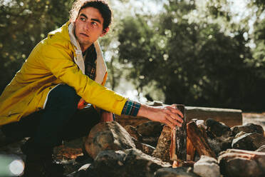 Side view of a young man camping in a countryside location setting up a bonfire. Man sitting on ground putting fire wood for a campfire in a forest. - JLPSF15874