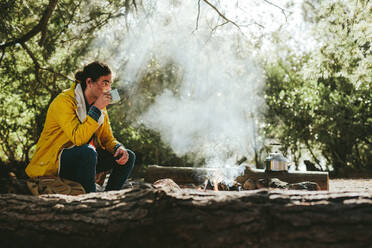 Mann genießt eine Tasse Tee an einem Lagerfeuer im Wald. Reisende auf einer Abenteuerreise auf dem Lande sitzen an einem Lagerfeuer und trinken Tee. - JLPSF15877