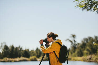 Seitenansicht eines Fotografen, der durch seine auf einem Stativ montierte DSLR-Kamera im Freien schaut. Ein Mann steht an einem See und fotografiert die Natur an einem sonnigen Tag. - JLPSF15882