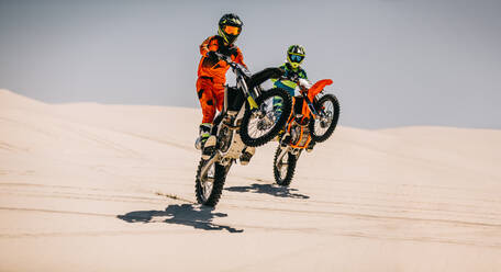 Two motocross riders doing wheelie together over sand dunes. Stunt bike riders riding motorcycle on one wheel in desert. - JLPSF15902