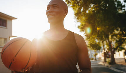 Nahaufnahme eines Sportlers, der auf der Straße steht und einen Basketball hält, mit Sonne im Hintergrund. - JLPSF15915