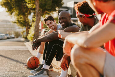 Vier Basketballspieler entspannen sich nach einem Basketballspiel auf dem Bürgersteig sitzend. Fröhliche Sportler sitzen auf dem Bürgersteig und diskutieren. - JLPSF15919