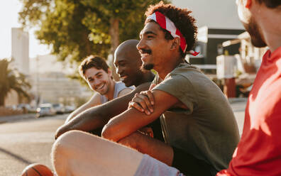 Nahaufnahme von vier Basketballern, die sich nach einem Basketballspiel auf dem Bürgersteig ausruhen. Fröhliche Sportler sitzen auf dem Bürgersteig und diskutieren. - JLPSF15920