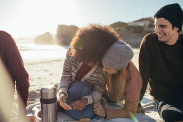 Two young woman sitting on the beach looking something funny on mobile phone and smiling with a friends sitting by. Group of young people relaxing on the sea shore. - JLPSF15927