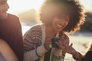 Junge Frau mit Kaffeetasse, die lachend mit Freunden am Strand sitzt. Fröhliche Freunde am Strand bei Sonnenuntergang. - JLPSF15938