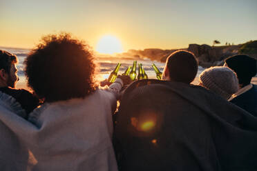 Group of friends cheers with beers at the beach. Young people sitting together at beach during sunset and having a party. - JLPSF15941