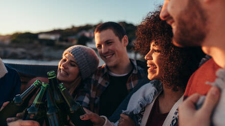 Eine Gruppe von Freunden feiert und trinkt am Abend am Strand. Junge Menschen verschiedener Rassen stoßen mit Bierflaschen am Strand an. - JLPSF15945