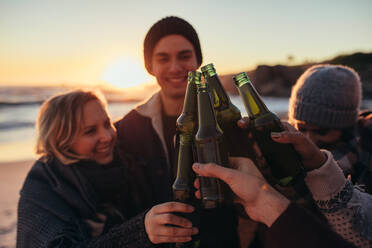 Junge Leute stoßen mit Bierflaschen am Strand an. Eine Gruppe von Männern und Frauen trinkt während des Sonnenuntergangs gemeinsam am Meeresufer. - JLPSF15946