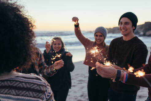 Eine bunt gemischte Gruppe junger Leute feiert den Neujahrstag am Strand. Junge Leute haben Spaß mit Wunderkerzen im Freien am Meeresufer. - JLPSF15956