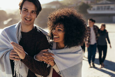 Portrait of a couple wrapped in a blanket while walking on a sea shore with friends at the back. Man and woman walking on the beach looking away and smiling. - JLPSF15959