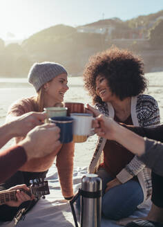 Eine Gruppe von Freunden feiert abends am Strand eine Party mit Kaffee. Junge Leute stoßen am Strand auf Kaffeetassen an. - JLPSF15972