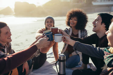 Group of multi-ethnic young people toasting coffee mugs on the beach. Group of friends spending time together at the beach having coffee. - JLPSF15974