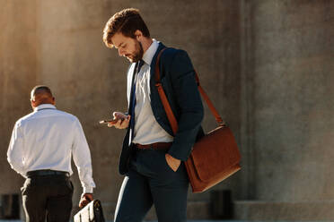 Men in formal clothes commuting to office early in the morning carrying office bags. Businessman using mobile phone while walking on city street to office. - JLPSF15987