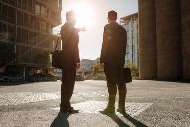 Rear view of business colleagues carrying office bags standing on street looking away with one person pointing towards a distant object. Business people standing on a street looking away while commuting to office with sun in the background. - JLPSF15993