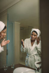 Afrikanische Frau im Bademantel, die kosmetische Lotion aufträgt, um die Haut im Badezimmer zu pflegen. Gesichtspflegekonzept. Reflexion der reifen Frau im Spiegel, die Gesichtskosmetik im Badezimmer aufträgt. Morgenhygiene. - JLPSF15998