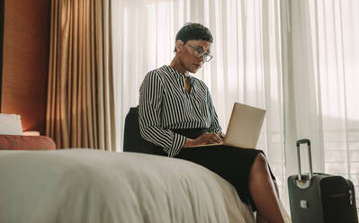 Geschäftsfrau sitzt auf dem Bett und benutzt einen Laptop. Afrikanische Frau auf Geschäftsreise arbeitet im Hotelzimmer. - JLPSF16000