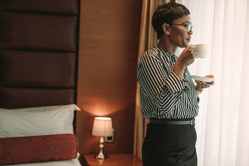 Mature african woman in formal clothes standing in hotel room and having a cup of coffee. Businesswoman standing in hotel room drinking coffee. - JLPSF16003