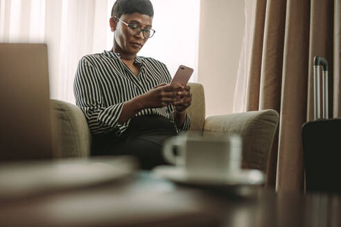 Reife afrikanische Geschäftsfrau sitzt auf Hotelzimmercouch und benutzt sein Handy. CEO auf Geschäftsreise schreibt SMS auf Handy im Hotelzimmer. - JLPSF16004