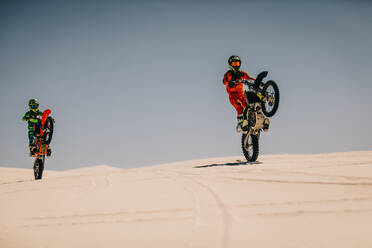 Two motocross racers performing stunt with motorcycles in desert. Dirt bike riders doing wheelie on sand dunes. - JLPSF16045