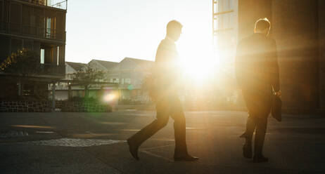 Männer auf dem Weg ins Büro am frühen Morgen mit der aufgehenden Sonne im Hintergrund. Geschäftsleute mit Bürotasche in der Hand gehen auf der Straße ins Büro. - JLPSF16049