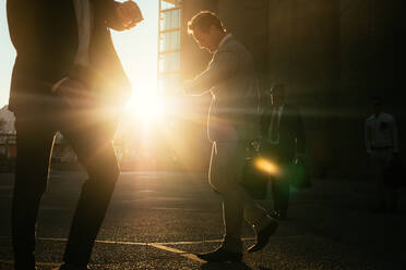 Männer auf dem Weg ins Büro am frühen Morgen, die Bürotaschen tragen und auf ihre Armbanduhr schauen. Geschäftsleute, die es eilig haben, ihr Büro zu erreichen, gehen auf der Straße mit der Sonne im Hintergrund. - JLPSF16051