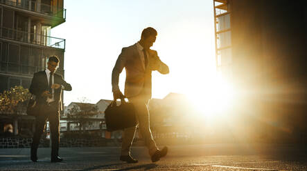 Men in formal clothes commuting to office early in the morning carrying office bags looking at their wrist watch. Businessmen in hurry to reach office walking on city street with sun flare in the background. - JLPSF16054