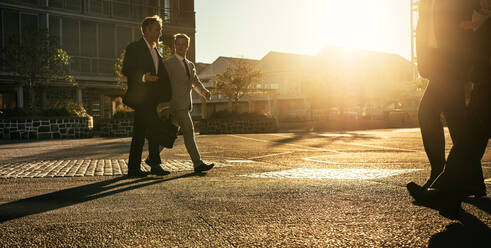 Männer in formeller Kleidung auf dem Weg ins Büro am frühen Morgen mit Bürotaschen. Geschäftskollegen im Gespräch beim Spaziergang auf der Straße in der Stadt mit Sonnenschein im Hintergrund. - JLPSF16056