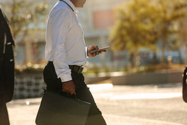 Businessman using mobile phone while commuting to office. Busy office going person walking on street carrying his office bag looking at his mobile phone. - JLPSF16064