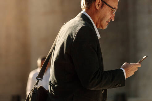 Man in formal clothes looking at his mobile phone while walking on street to office. Busy office going person walking on street and using his mobile phone. - JLPSF16072