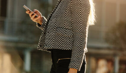 Mid section of a woman using her mobile phone while commuting to office. Busy office going woman walking on street and using her mobile phone with sun flare in the background. - JLPSF16075