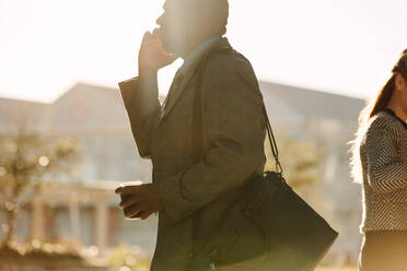 Geschäftsmann auf dem Weg ins Büro im Gespräch über Handy mit einer Kaffeetasse in der Hand mit Sonne Flare im Hintergrund. Busy Mann zu Fuß ins Büro trägt seine Bürotasche halten und eine Kaffeetasse in der einen Hand und im Gespräch über Handy mit der anderen. - JLPSF16077