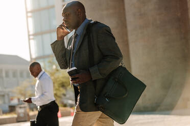Männer, die auf dem Weg ins Büro ein Mobiltelefon benutzen. Geschäftsleute, die ein geschäftiges Leben führen, benutzen Mobiltelefone und trinken Kaffee, während sie auf der Straße ins Büro gehen. - JLPSF16082