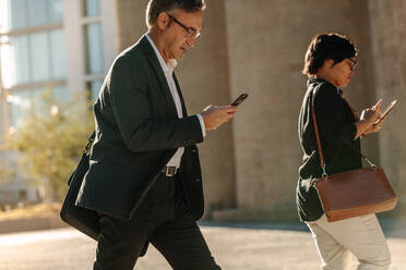 Ein Mann und eine Frau benutzen ihr Handy auf dem Weg ins Büro auf einer Straße in der Stadt. Geschäftsleute überprüfen ihr Handy auf dem Weg ins Büro und zeigen ihren vollen Terminkalender. - JLPSF16085