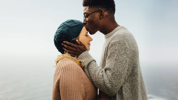 Seitenansicht eines liebenden gemischtrassigen Paares im Freien. Junger Mann küsst die Stirn seiner Freundin. - JLPSF16091