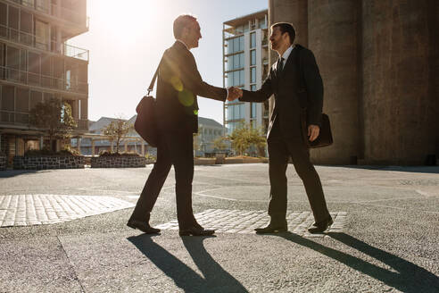 Business colleagues greeting each other and shakings hands on a street while commuting to office. Side view of businessman shaking hand with a colleague outdoors with sun in the background. - JLPSF16092