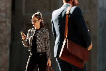 Geschäftsfrau auf dem morgendlichen Weg ins Büro, die auf ihr Mobiltelefon schaut. Geschäftsleute, die auf der Straße ins Büro gehen und ihre Bürotaschen tragen und dabei ihr Mobiltelefon benutzen. - JLPSF16094