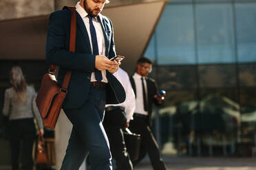 Businessman in formal clothes carrying office bag using mobile phone while commuting to office. Busy office going people using their mobile phones while walking on street. - JLPSF16098