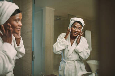 Reife Frau im Bademantel, die in den Badezimmerspiegel schaut, während sie Feuchtigkeitscreme auf ihr Gesicht aufträgt. Afrikanische Frau, die Creme auf das Gesicht im Badezimmer zu Hause aufträgt. - JLPSF16104