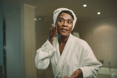 Reflection of african woman in bathroom mirror applying cosmetic cream on her face. Mature female putting on moisturizer on her face skin. - JLPSF16105