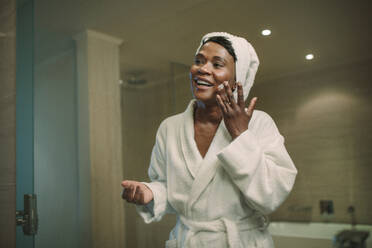 Smiling african woman applying cosmetic cream on face standing in the bathroom. Reflection in mirror of female putting body lotion cream on her face. - JLPSF16106