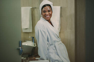 Porträt einer reifen afrikanischen Frau mit glänzender Haut, die im Bademantel an einem Waschbecken lehnt und lächelnd in die Kamera schaut. - JLPSF16111