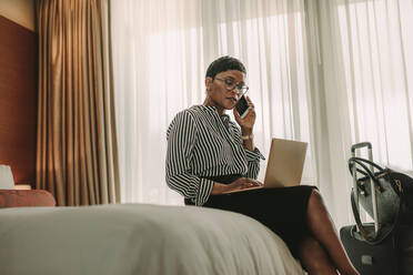 Reife afrikanische Geschäftsfrau arbeitet mit Laptop und Mobiltelefon im Hotelzimmer. Weiblicher CEO auf Geschäftsreise arbeitet vom Hotelzimmer aus. - JLPSF16116