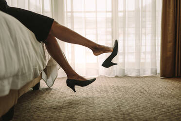 Professionelle Business-Frau entspannt sich auf dem Bett eines Hotelzimmers, nachdem sie zur Arbeit gereist ist. Eine Frau, die nach einer langen Reise müde ist und sich auf dem Bett ausruht. Fokus auf eine Frau, die ihre Schuhe auszieht und auszieht, während sie im Bett sitzt. - JLPSF16119