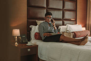 Frau in formeller Kleidung sitzt auf dem Bett und spricht mit einer Zeitung in der Hand über ein Mobiltelefon. Afrikanische Geschäftsfrau in einem modernen Hotelzimmer, die eine Zeitung liest und einen Anruf tätigt. - JLPSF16123